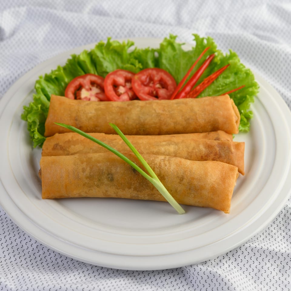 Golden-brown spring rolls on a white plate, garnished with lettuce, tomato slices, red chilies, and green onions.